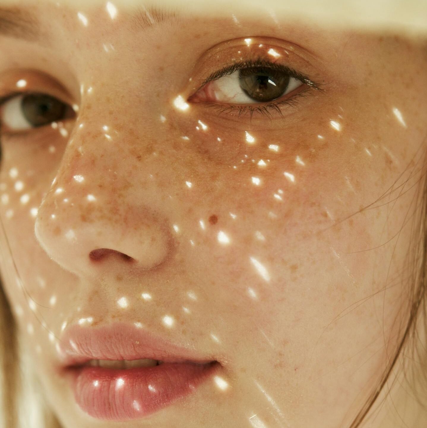 Close-up of a woman with freckles wearing a hat, sunlight creating patterns on her face.